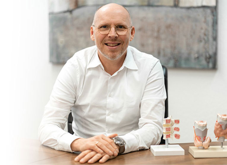 Prof. Dr. Herrmann – Endokrinologie & Diabetologie in Bochum Prof. Dr. Herrmann mit Brille in weißem Hemd sitzt an einem Tisch mit medizinischen Modellen im Hintergrund